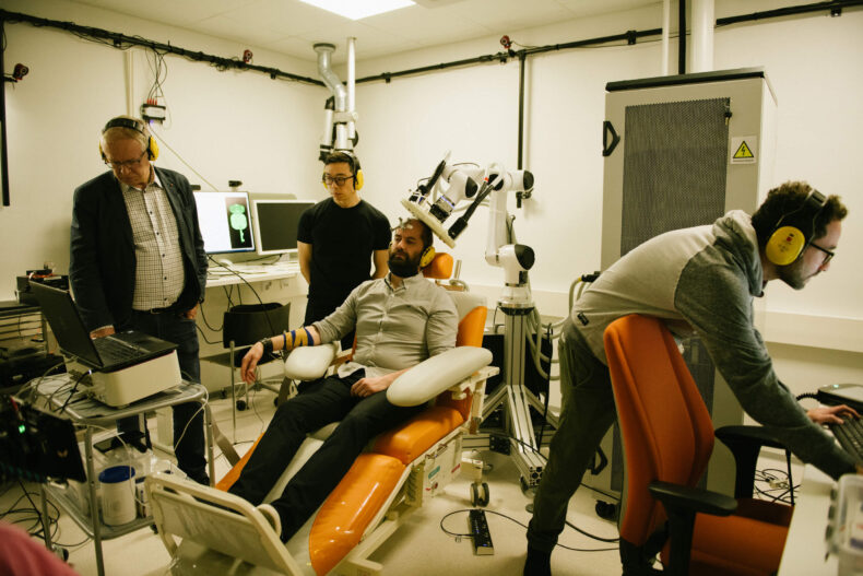 The image featores one person sitting in a medical recliner chair, with three other people standing around them. Everyone is wearing ear protection. The sitting person has a medical device touching their head.